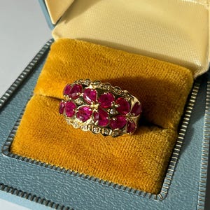 May include: A gold ring featuring a floral pattern, adorned with bright red gemstones and tiny diamonds. The ring is presented in a vintage blue jewellery box with a yellow velvet lining, offering a luxurious display.
