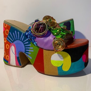 May include: A pair of colorful platform sandals with a multi-colored geometric design. The sandals have a thick platform sole and are decorated with a large purple gem, green sequins, and gold wire.
