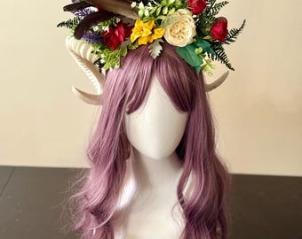 May include: A decorative headpiece featuring a floral crown with red, yellow, and cream-colored flowers, green leaves, and dark feathers. The headpiece is adorned with curved, off-white horns and rests on a mannequin head with long, wavy, purple hair.