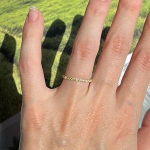 Könnte beinhalten: Ein zarter Goldring mit kleinen, klaren Edelsteinen, die in Pavé-Fassung gefasst sind. Der Ring wird am Ringfinger einer Hand mit manikürten Nägeln getragen.