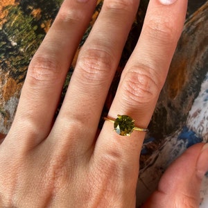 May include: A gold ring with a round, green gemstone. The ring is on a person's finger.