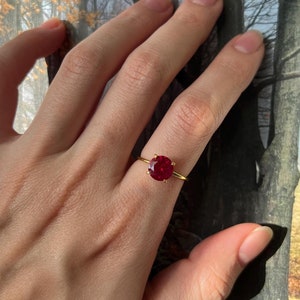 May include: A gold ring with a large, round, red gemstone. The ring is on a person's finger.