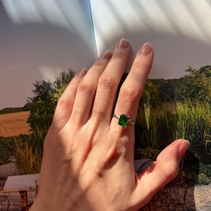 Puede incluir: Un anillo de plata con una piedra cuadrada verde esmeralda. El anillo est&aacute; en el dedo de una persona.
