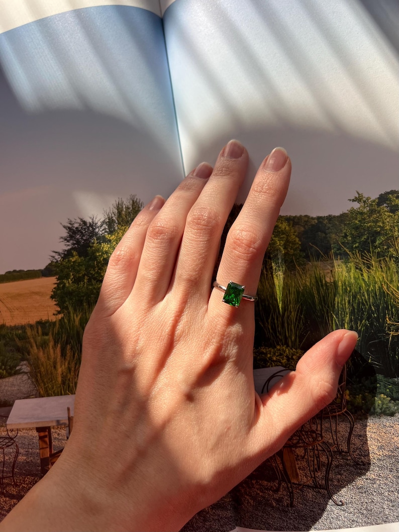 Puede incluir: Un anillo de plata con una piedra cuadrada verde esmeralda. El anillo est&aacute; en el dedo de una persona.