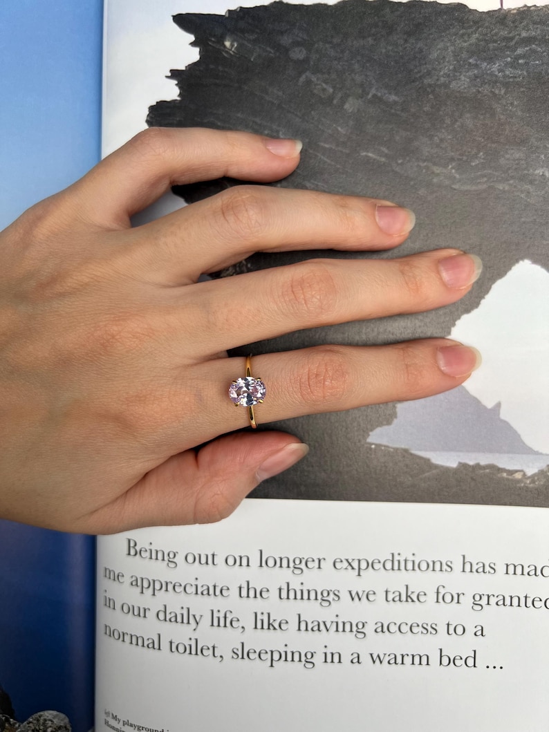 May include: A gold ring with a light pink oval-shaped gemstone. The ring is on a finger.