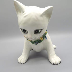 May include: White ceramic kitten figurine with green, yellow, and blue flower accents around its neck. The kitten is sitting with its head down and its paws tucked under its body.