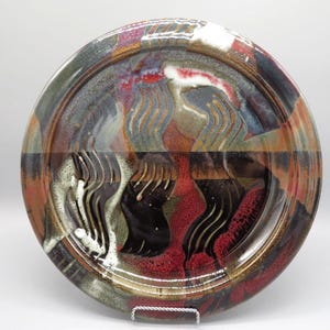 May include: A handcrafted ceramic plate with a unique, abstract design. The plate features a blend of colors, including red, black, white, and shades of brown and gray. The rim has a dark border, and the center showcases a textured, wave-like pattern.