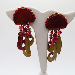 May include: A pair of statement earrings featuring a deep red, half-circle design with a beaded border. Dangling from the base are various red beads and brass-colored, leaf-shaped charms. The earrings are displayed on a white stand.