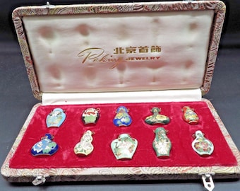 May include: An open jewelry box with a red velvet interior displays ten small, colorful, decorative perfume bottles. The bottles are various shapes and feature intricate designs in blue, green, red, and gold. The box is labeled "Peking Jewelry".