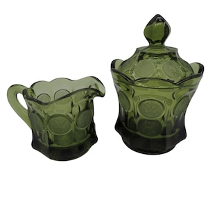 May include: A green glass creamer and sugar bowl set with a coin design. The creamer has a handle and a scalloped rim. The sugar bowl has a lid with a finial.