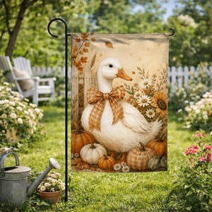 May include: A rectangular garden flag featuring a white duck wearing a brown plaid bow around its neck. The duck is surrounded by orange and white pumpkins, autumn leaves, and white daisies against a light beige background with a wooden fence.