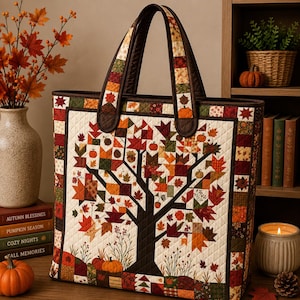 May include: A quilted tote bag featuring a central dark brown tree silhouette against a cream background, adorned with colorful fabric patches of autumn leaves, pumpkins, and acorns. The bag has two dark brown handles and a patchwork border.