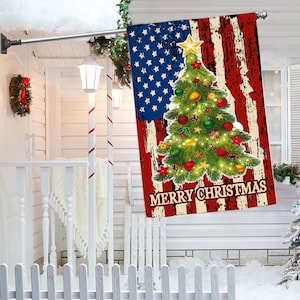 May include: A Christmas flag featuring the American flag design with a decorated Christmas tree in the center. The flag has the words "MERRY CHRISTMAS" at the bottom. The flag is hanging on a porch.