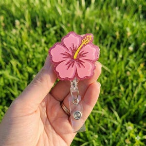 May include: A pink hibiscus flower badge reel with a glittery outline and yellow stamens. The retractable reel has a clear plastic strap and a metal clip. The background is blurred green grass.