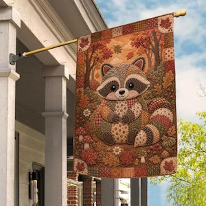 May include: A rectangular garden flag featuring a patchwork-style illustration of a raccoon sitting in an autumn forest. The design includes brown, orange, and red fall leaves, mushrooms, and acorns, with a checkered border on a gold metal pole.