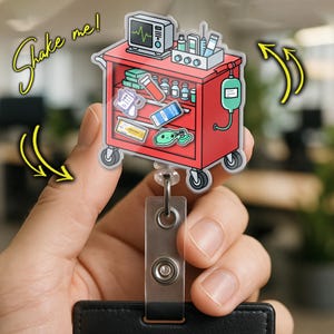 May include: A red medical cart badge reel with various medical supplies and equipment. The badge reel has a clear plastic front and a retractable cord. The words "Shake me!" are written in yellow script. The badge reel is held by a person.