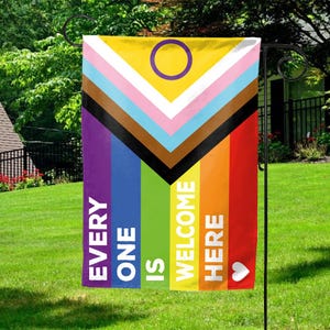 May include: A garden flag with a colorful design. The flag features a yellow top with a purple circle, and a series of stripes in various colors, including pink, blue, brown, and black. The flag also has the words "EVERY ONE IS WELCOME HERE" in white text, with a white heart.