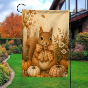 May include: A rectangular garden flag featuring a brown squirrel wearing a tan and white plaid bow tie. The squirrel is surrounded by orange and white pumpkins, white daisies, and a wooden fence against a backdrop of autumn leaves.