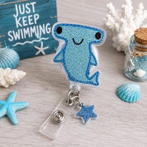 May include: A blue glitter hammerhead shark badge reel with a smiling face and a blue glitter star charm. The reel has a clear plastic strap and a metal clip. The background includes a wooden sign that says "JUST KEEP SWIMMING".