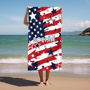 May include: A beach towel with a patriotic design featuring red and white stripes, blue stars, and the name "SOPHIA" in white. The towel is held up on a beach with the ocean in the background.