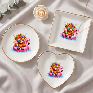 May include: Three white ceramic trinket dishes with gold-toned rims, featuring a graphic of a brown highland cow wearing rainbow sunglasses and a flower crown while floating on a pink flamingo pool float and holding a colorful drink.