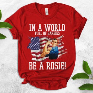 May include: Red t-shirt with a graphic of a woman in a blue jumpsuit holding a wrench in front of an American flag. The text "In a world full of Barbies be a Rosie!" is printed on the shirt.
