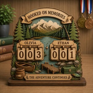 May include: A wooden layered display featuring a mountain lake scene with the text HOOKED ON MEMORIES and THE ADVENTURE CONTINUES. It includes two sets of flip-style number counters labeled OLIVIA and ETHAN, a small boat, a dock, and a tackle box.