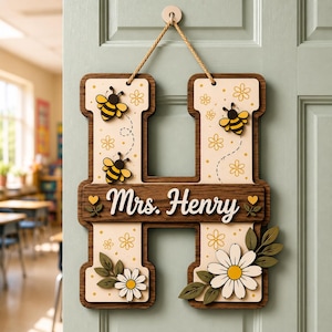 May include: A wooden H-shaped wall sign featuring a cream-colored background with yellow flower patterns, three small bees, and two white daisies with green leaves. The center horizontal bar displays the white text Mrs. Henry with two small yellow hearts.