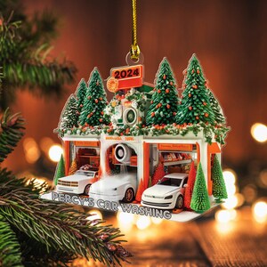 May include: A Christmas ornament shaped like a car wash, featuring three white cars. The ornament has a sign that reads "2024" and "Percy's Car Washing". The car wash is decorated with miniature Christmas trees and garlands.