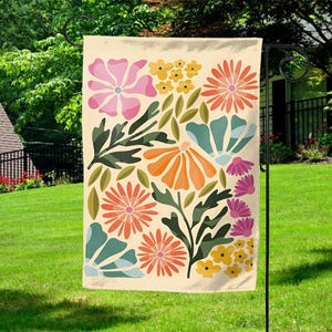 May include: A beige garden flag featuring a colorful floral design. The design includes pink, orange, yellow, and teal flowers, along with green leaves. The flag is hanging on a black metal stand in a grassy yard.