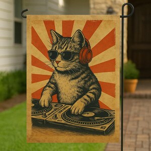 May include: A garden flag featuring a tabby cat wearing sunglasses and orange headphones, DJing with turntables. The background has a retro orange and cream sunburst design. The flag is rectangular and hangs from a black metal pole.