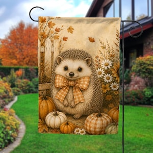 May include: A rectangular garden flag featuring a central illustration of a hedgehog wearing a large orange and white plaid bow. The hedgehog is surrounded by several pumpkins, white daisies, and a wooden fence, all set against a warm autumn background.