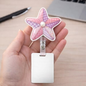 May include: A retractable badge holder with a pink and purple patchwork starfish design, a white border, and a pearl accent. The holder has a clear plastic clip and a blank white rectangular badge.
