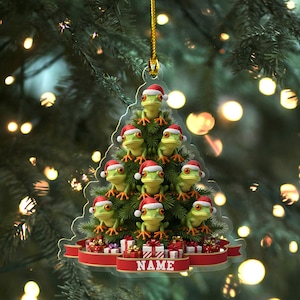May include: A Christmas tree ornament shaped like a Christmas tree, featuring green frogs wearing Santa hats. The ornament has a red ribbon base with the word "NAME" printed on it, and small gift boxes. The ornament is hanging from a gold string.