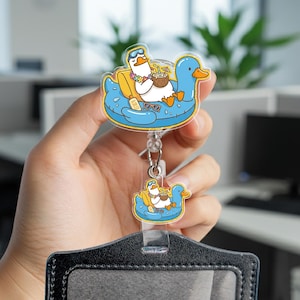 May include: A retractable badge reel featuring two layered acrylic charms of a white duck wearing goggles and a floral lei, relaxing in a blue duck-shaped pool float with a coconut drink, sunglasses, and a bottle labeled SPF 50, attached to a black holder.