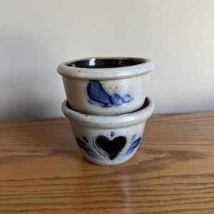 May include: Two stacked, small, stoneware pots with a speckled, light gray finish. The top pot has a dark blue design, and the bottom pot features a heart-shaped cutout and blue leaf accents. The interior of the pots is dark.
