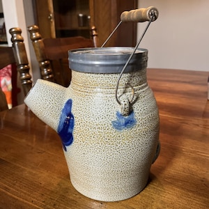 May include: A vintage stoneware pitcher with a spout and a metal handle. The pitcher is speckled with a tan and brown glaze, and features blue floral accents. The handle has a wooden grip.