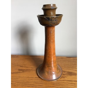 May include: A tall, glazed ceramic candlestick holder with a flared base and a cylindrical stem. The lower portion features a warm, mottled orange-brown finish, while the upper candle cup is a darker, brownish-grey with visible crackled texture.