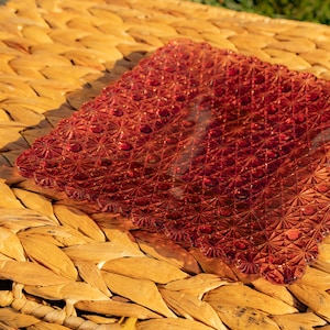 Puede incluir: Un plato cuadrado de vidrio rojo rubí con un patrón geométrico texturizado. El plato tiene un borde festoneado y se asienta sobre una superficie tejida de color paja. El diseño del plato presenta un patrón repetido de pequeños círculos y formas de estrella.