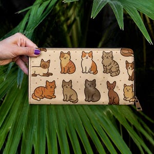 May include: A beige wallet with a repeating pattern of cartoon cats in different colours and poses. The wallet has a zip closure. It is held against a backdrop of green foliage.