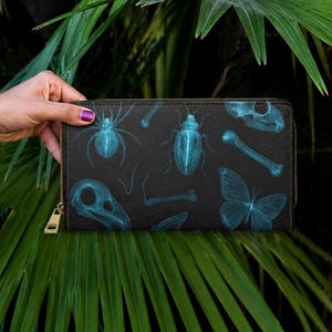 May include: Black zip-around wallet with teal illustrations of insects, bones, and a butterfly. The wallet is held against green palm leaves. The zip pull is gold.
