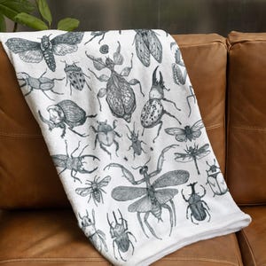 May include: A white throw blanket featuring a repeating black ink illustration pattern of various insects, including beetles, a praying mantis, a dragonfly, and a moth, displayed in a detailed, scientific sketch style across the fabric surface.