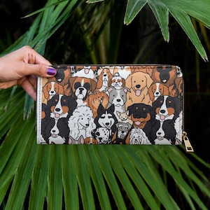 Könnte beinhalten: Eine rechteckige Geldbörse mit einem farbenfrohen Muster verschiedener Hunderassen. Die Geldbörse hat einen goldenen Reißverschluss und wird vor einem Hintergrund aus grünem Laub gehalten. Das Design umfasst Rassen wie Golden Retriever und Dalmatiner.