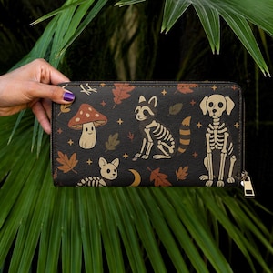 May include: Black wallet with a zip closure, decorated with a pattern of a mushroom, skeleton raccoon, skeleton dog, leaves, and a small dog skeleton. The wallet is set against green foliage.