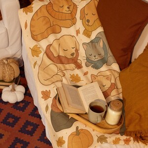 Könnte beinhalten: Eine beige Decke mit Cartoon-Katzen und -Hunden mit Schals, umgeben von Herbstblättern. Ein Holztablett enthält ein Buch, eine Tasse Tee und eine Kerze. Kürbisse und Kissen vervollständigen die gemütliche Szene.