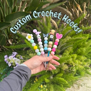 May include: Five colorful crochet hooks with personalized beads. The beads feature a flower, dinosaur, butterfly, cow, and heart. The hooks are labeled with the letters N, J, and L.