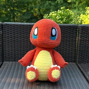 May include: A handmade crochet plush toy in the shape of a Charmander. The toy is primarily orange with a yellow belly and white accents on the feet. It has large, expressive eyes with white and blue details. The plush is sitting on a brown wicker surface.