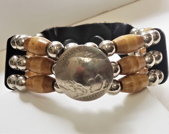 Può includere: Un braccialetto in pelle marrone con perline d'argento e perline di legno. Una moneta d'argento con la scritta "UNITED STATES OF AMERICA FIVE CENTS" è centrata sul braccialetto.