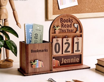 May include: A wooden desk organizer with a bookmark holder and a counter that reads "Books Read This Year". The counter displays "021" and the name "Jennie". The organizer has a bookshelf design.