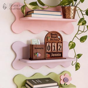 May include: Three-tiered, wavy-edged shelves in pink and green hold books, a potted plant, and decorative wooden accessories. A wooden calendar displays "Books Read This Year" and the number "012" with the name "Jennie". A wooden box reads "Bookmarks".